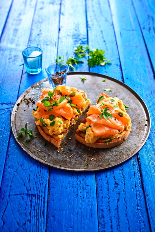 Zwei Scheiben geröstetes Brot mit cremigem Rührei und zartem Lachs auf einem Teller serviert, dekoriert mit frischen Kräutern.