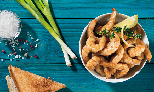 Ein Teller mit Garnelen, garniert mit Zitronenscheiben und Kräutern, auf einem blauen Holztisch mit Frühlingszwiebeln, Salz, Pfeffer und Toast.