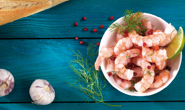 Schüssel mit Garnelensalat, garniert mit Dill, roten Pfefferkörnern und Limettenscheibe, auf blauem Holztisch. Knoblauch und Baguette im Hintergrund.
