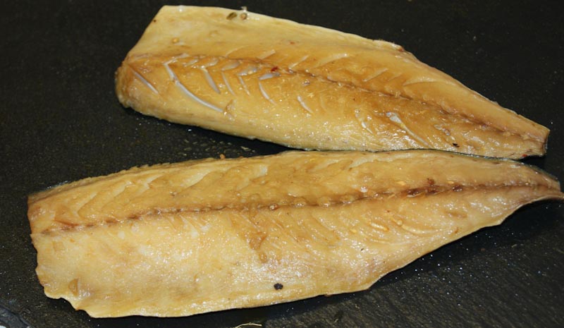 Zwei geräucherte Makrelenfilets auf einer dunklen Oberfläche liegend, die goldbraun glänzen und eine aromatische Textur haben.