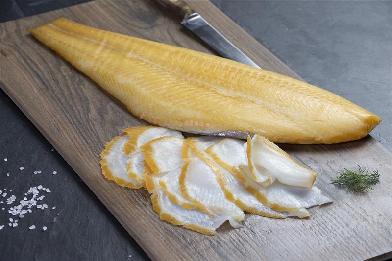 Ein großes Stück geräucherter Fisch liegt auf einem Holzschneidebrett, daneben sind dünne Scheiben des Fisches und ein Zweig Dill.