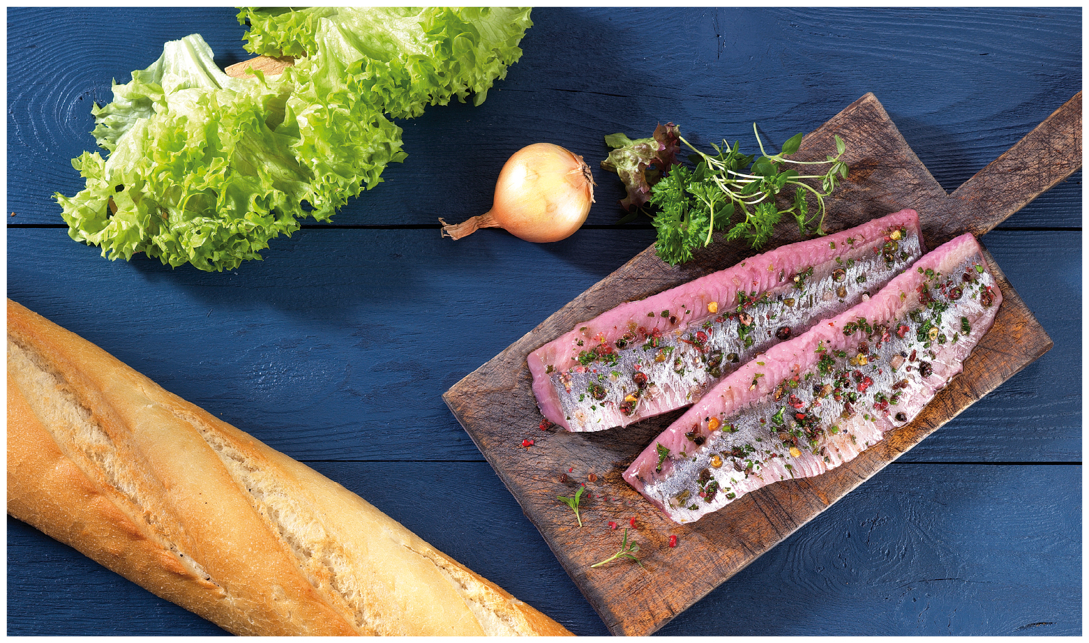 Heringsfilet auf Holzbrett, garniert mit Kräutern und Gewürzen, daneben Baguette, Salat und Zwiebel auf blauem Untergrund