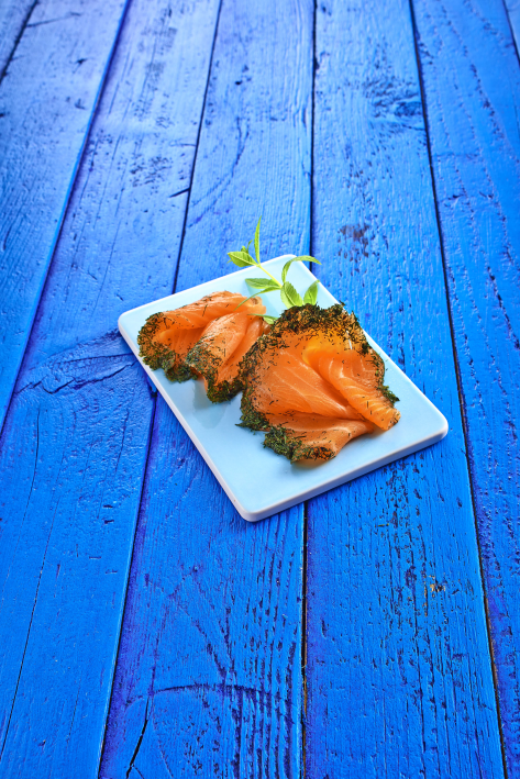 Geräucherter Lachs mit Kräutern auf weißem Teller Fein geschnittener, kräuterumrandeter Räucherlachs auf rechteckigem weißen Teller, auf blauem Holztisch serviert.
