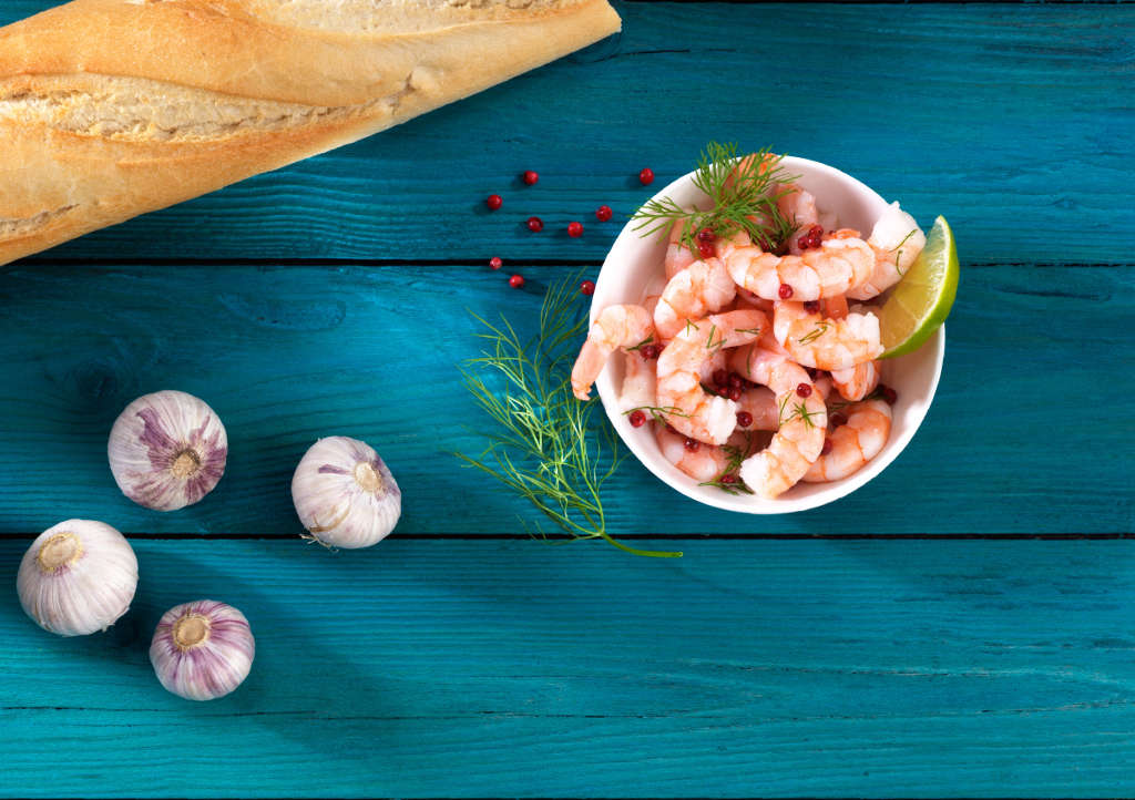 Eine Schale mit Garnelen, garniert mit Dill und rosa Pfefferkörnern, Limettenscheibe, daneben Baguette und Knoblauch auf blauem Holztisch